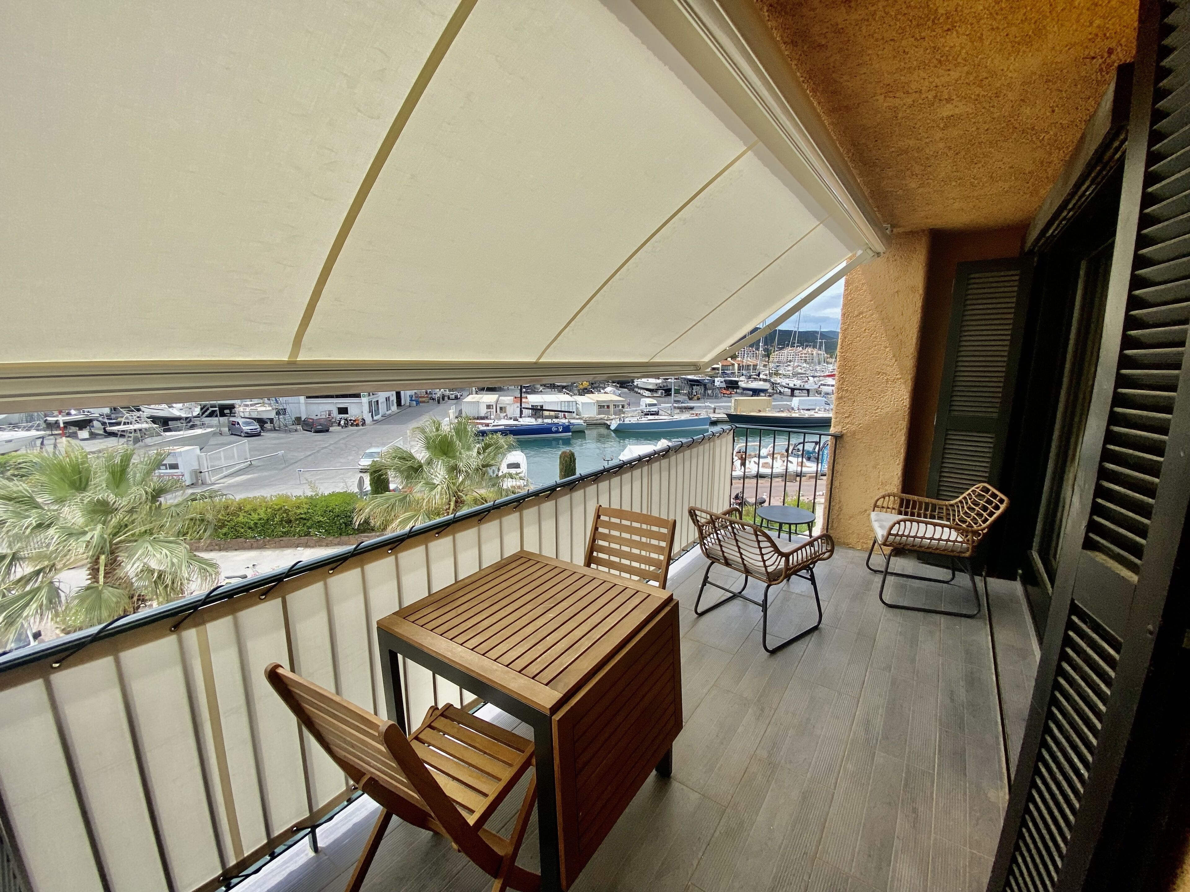 En balkon med et træbord og stole, udsigt over en lystbådehavn med både, og et palmetræ.