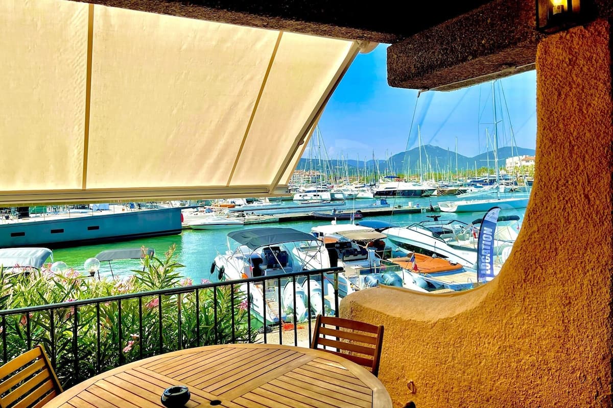 A terrace with a view of a marina, featuring boats and a mountainous backdrop.