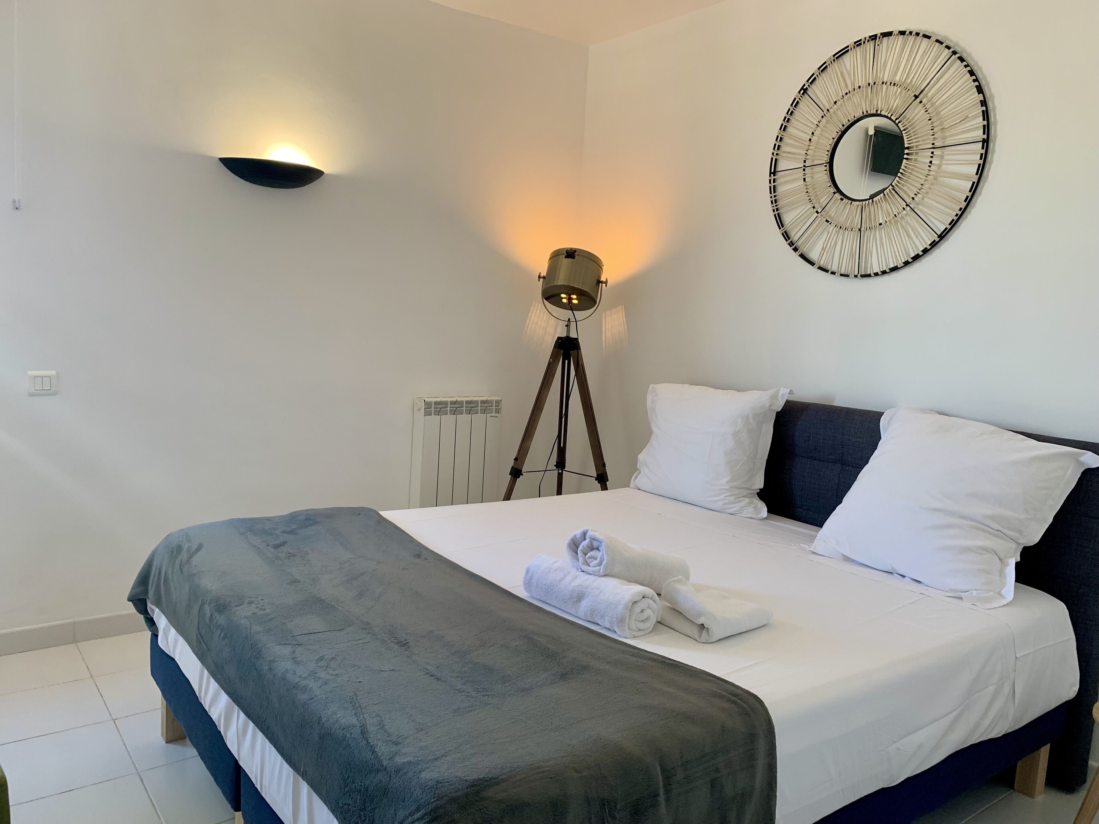Een keurig opgemaakt bed met wit en grijs beddengoed, een ronde wandklok en een vloerlamp.