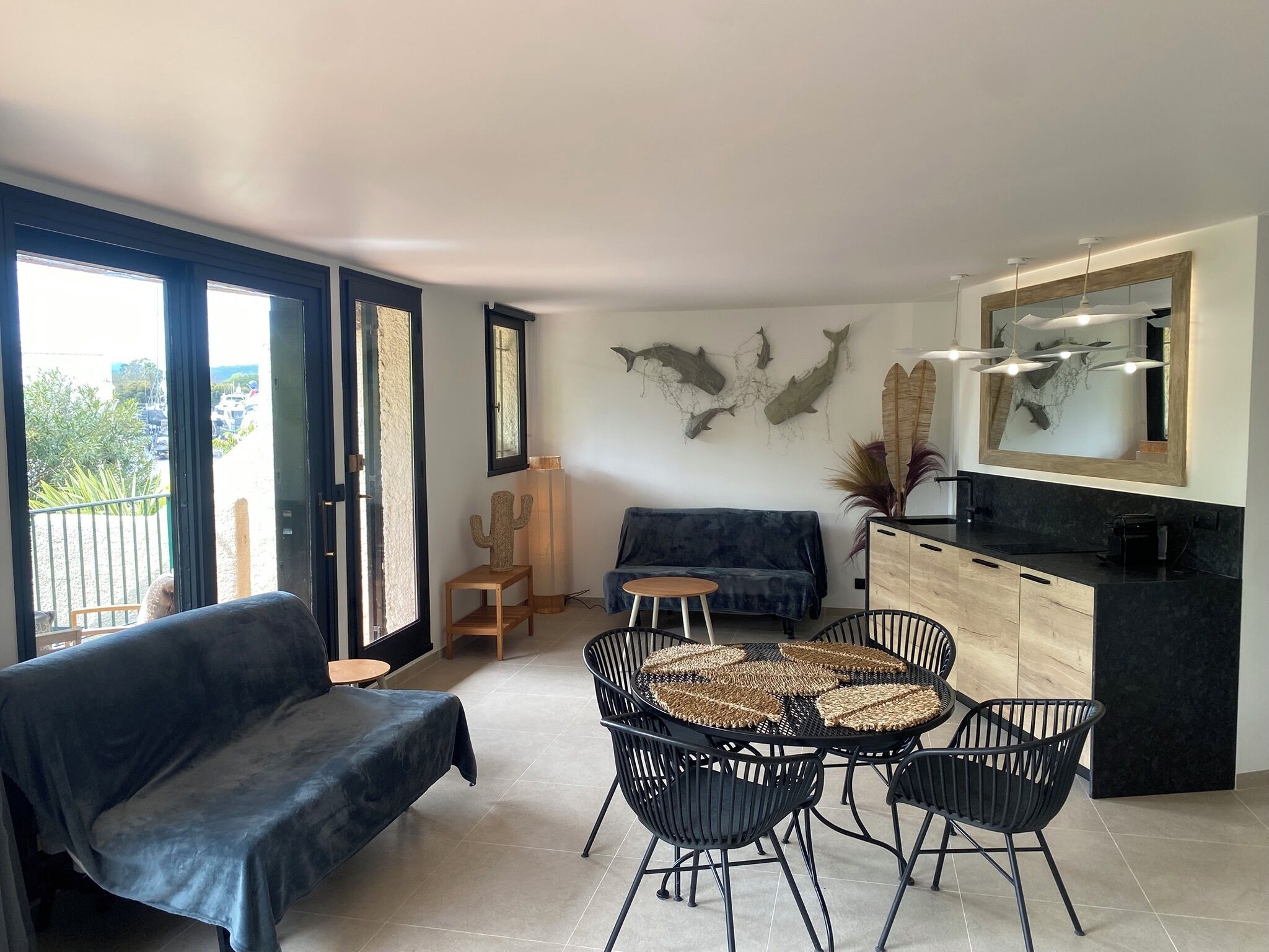 A modern living space with a sofa, a round dining table with chairs, a kitchen area with a countertop and cabinets, and a large window with a view of greenery.