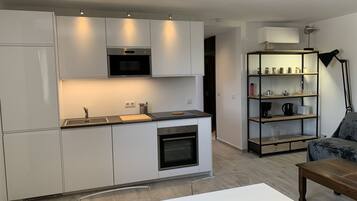 Una cocina moderna con armarios blancos, horno empotrado y microondas. En la encimera hay una tabla de cortar de madera, y una estantería con diversos objetos.