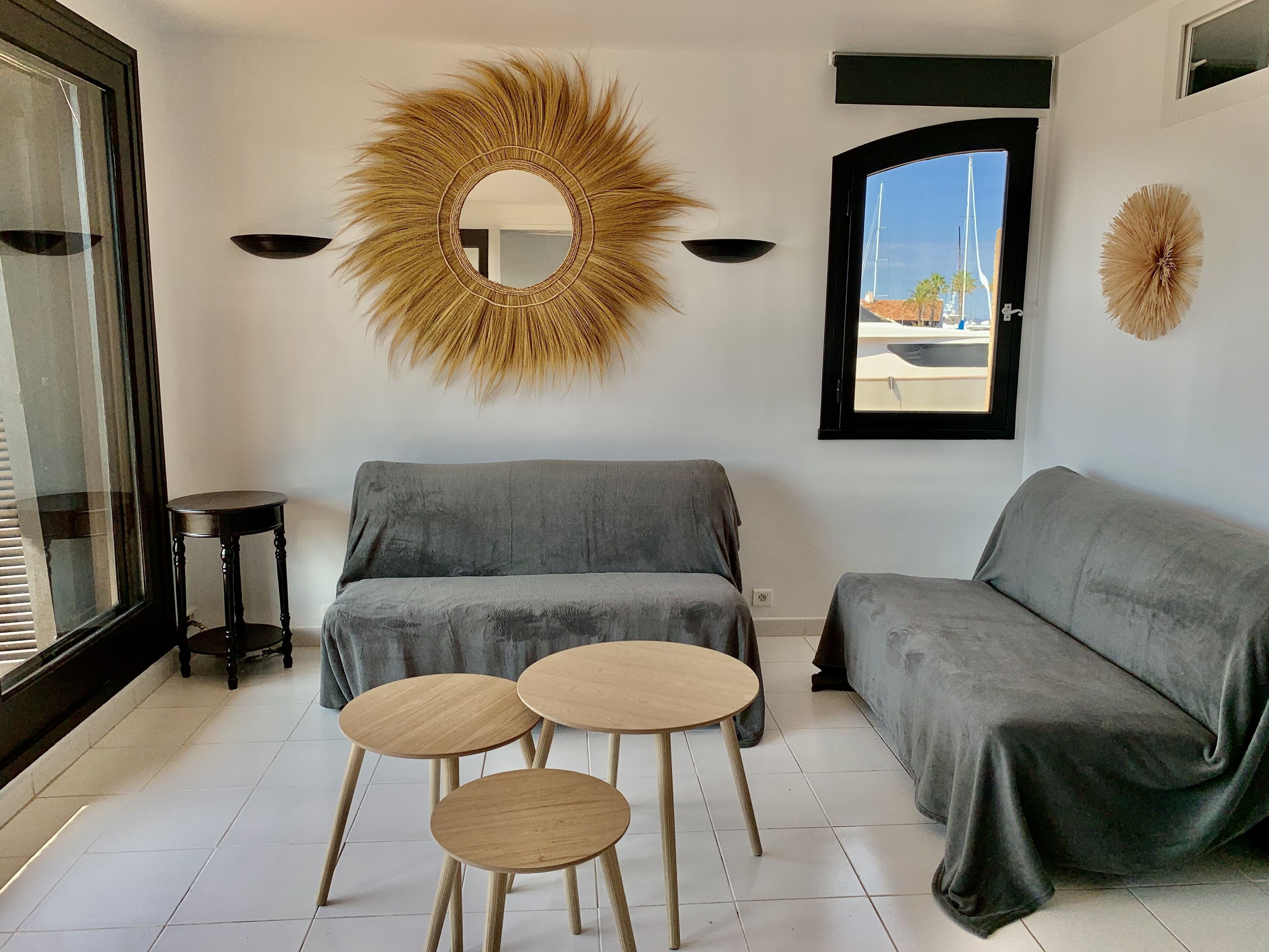A modern living room with a grey sofa, two round wooden tables, a large mirror, and a window with a view of boats.