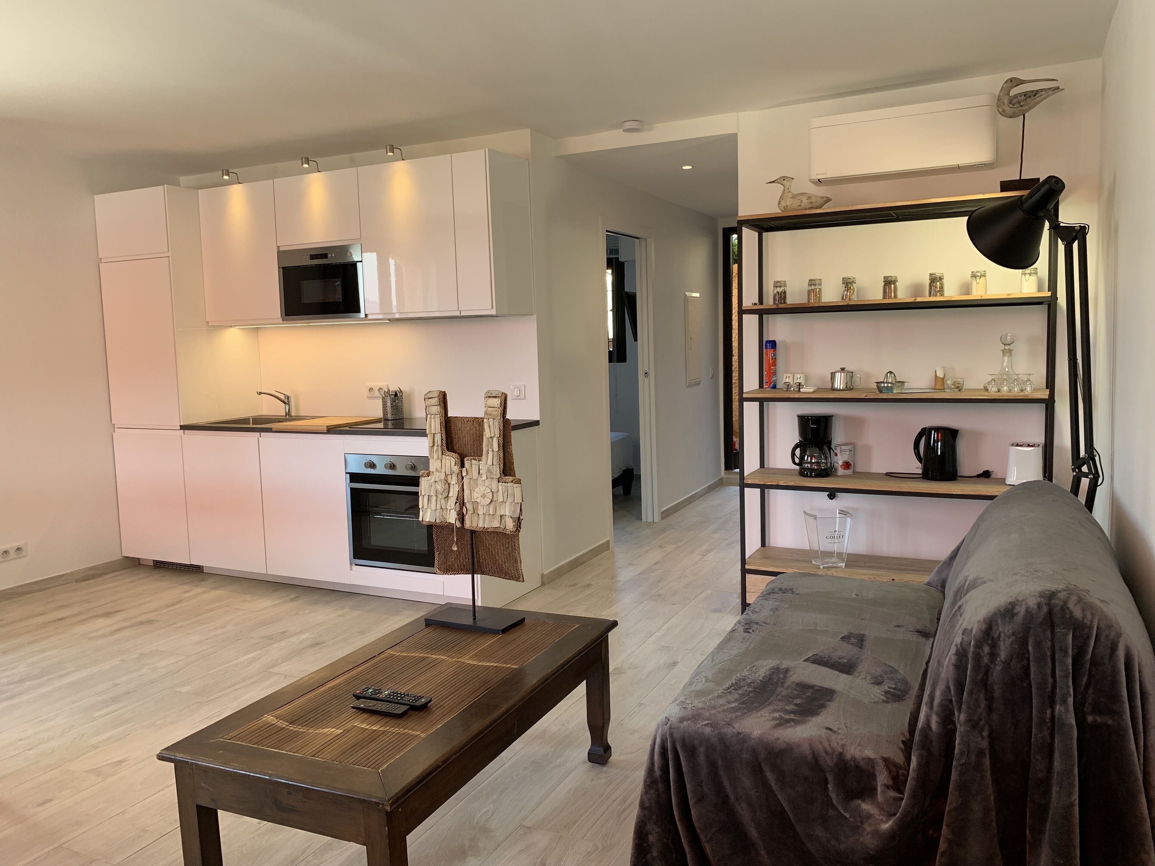 A modern kitchen with white cabinets, a microwave, and an oven. There is a wooden coffee table with a remote control. A cozy living area with a sofa and a bookshelf is visible.