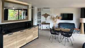Una cocina moderna con una isla grande, un comedor con sillas de mimbre, un televisor de pantalla plana y un ventanal con vistas a árboles.