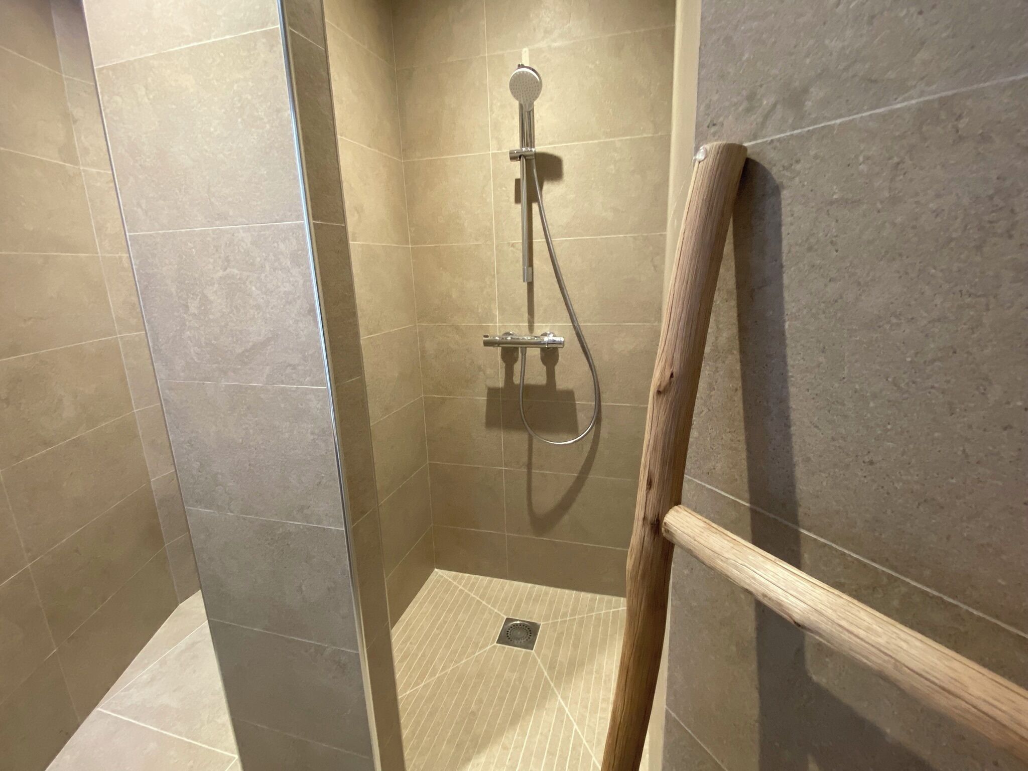 A shower area with a wooden handrail, tiled walls, and a drain in the floor.