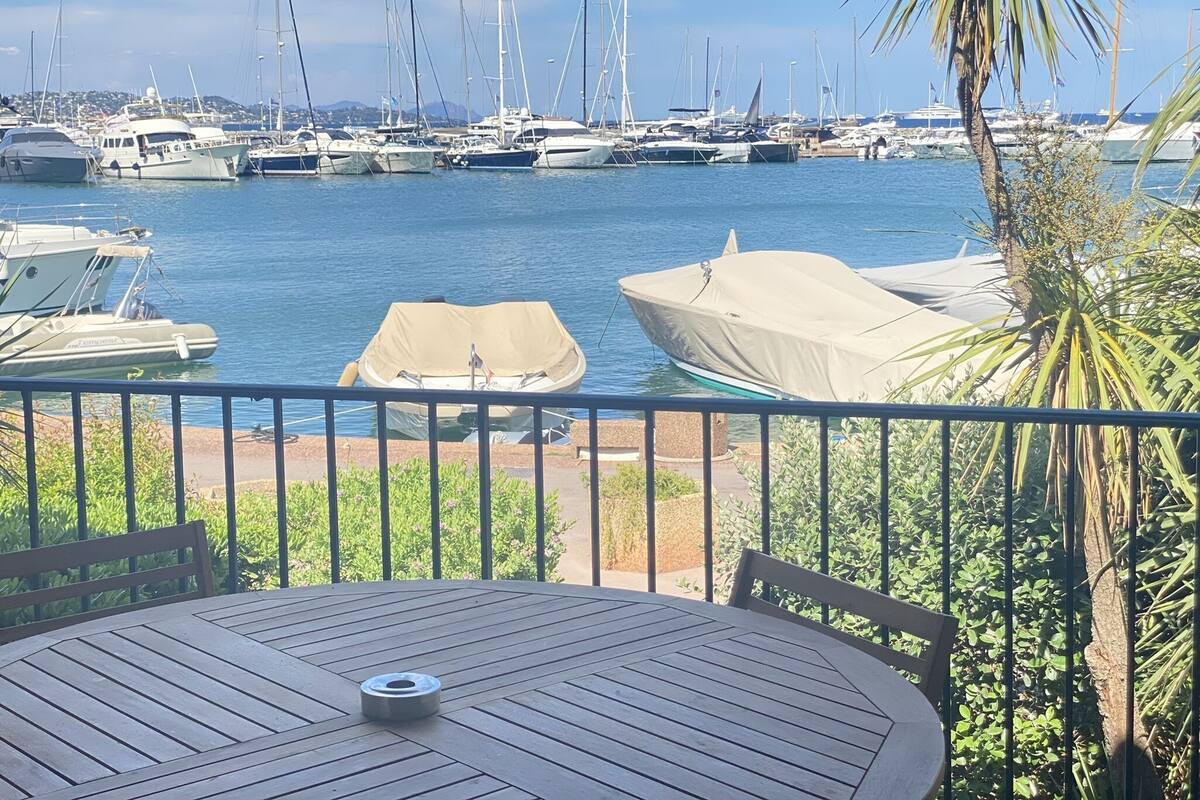 A marina with boats docked, a covered boat in the foreground, and a table with chairs on a balcony overlooking the water.