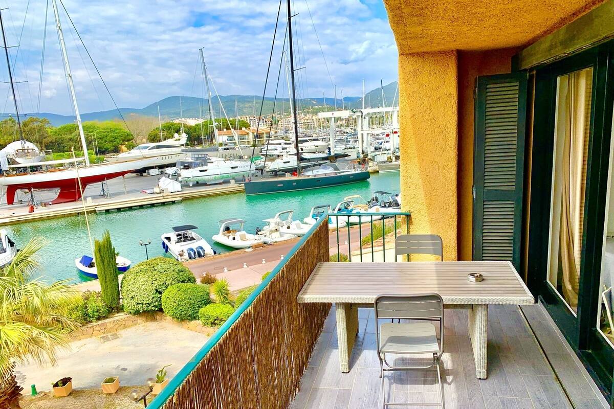 A balcony with a table and chairs overlooking a marina with boats and a mountainous landscape.