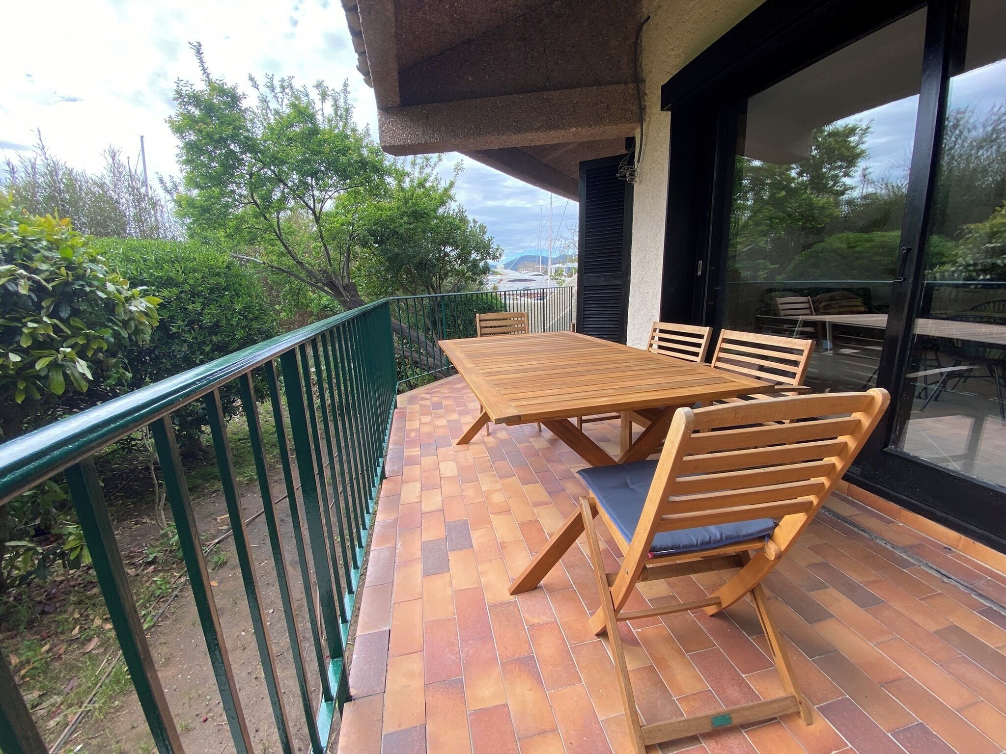 Una mesa y sillas de madera en un patio de ladrillo con barandilla verde, con vistas a un jardín y un edificio con amplias ventanas.