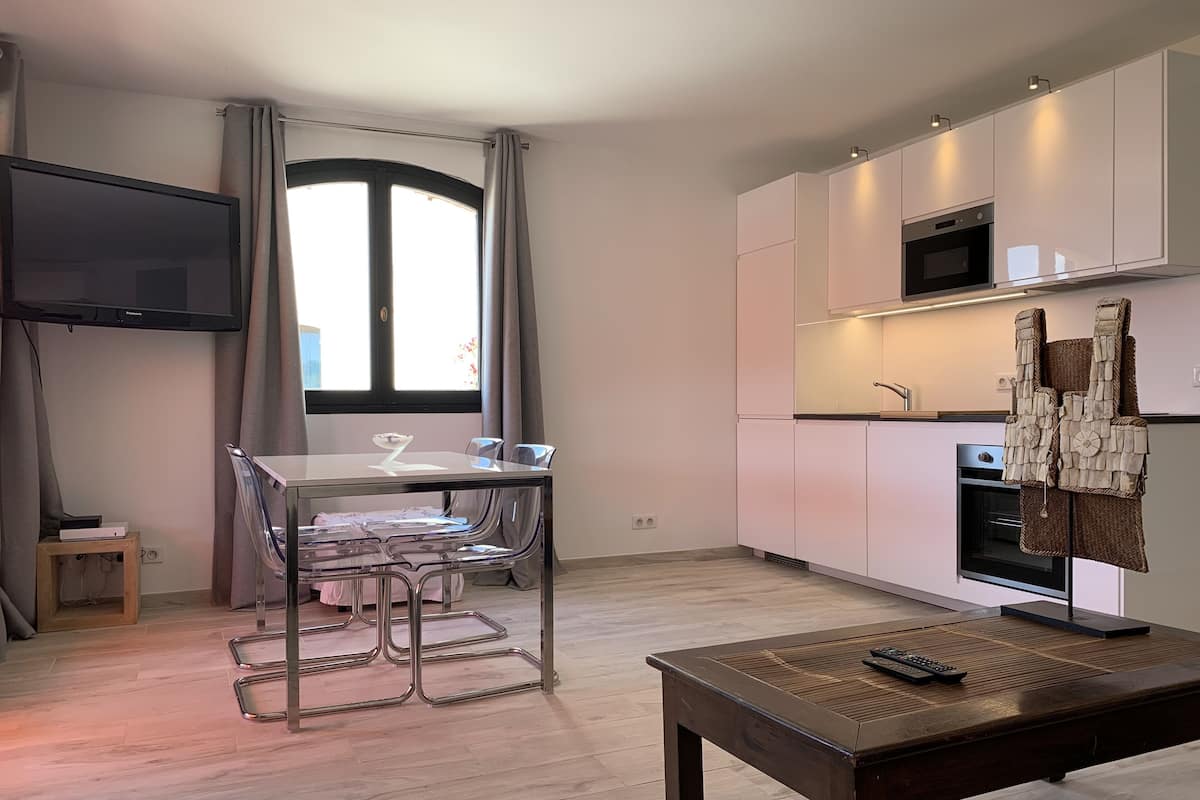 A modern kitchen with white cabinets, a microwave, and a built-in oven. There is a dining area with a glass table and chairs, and a TV mounted on the wall.