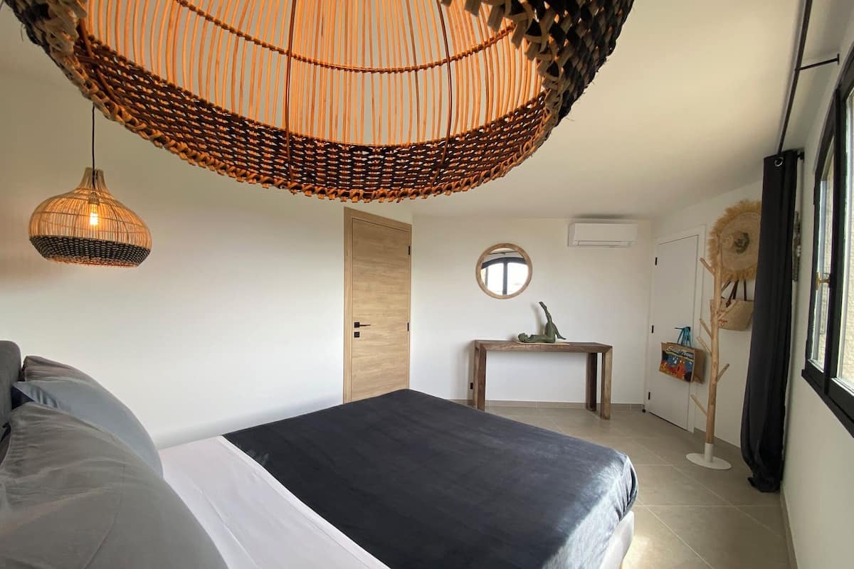 A bedroom with a bed, a wooden door, a small table with a statue, and a wicker hanging light fixture.
