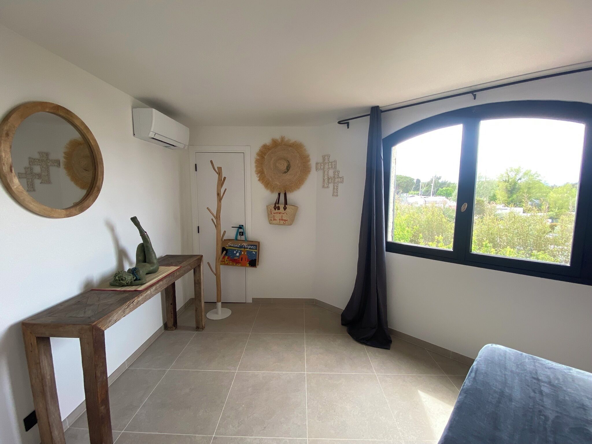 Una habitación con una mesa de madera, un espejo, aire acondicionado instalado en la pared, una puerta y una ventana con vistas a zonas verdes.