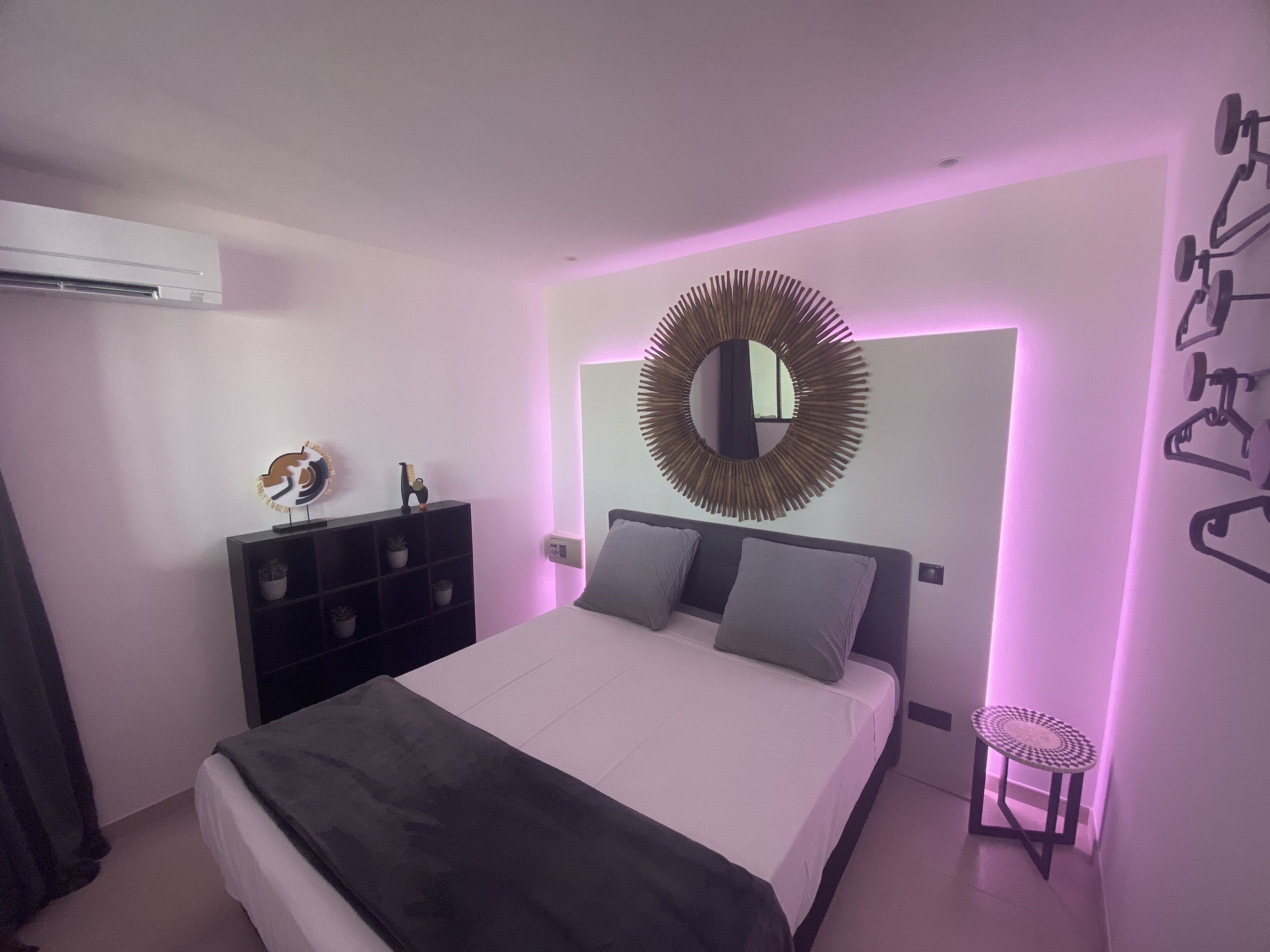A modern bedroom with a bed, a round mirror, a small stool, and a wall-mounted air conditioner.
