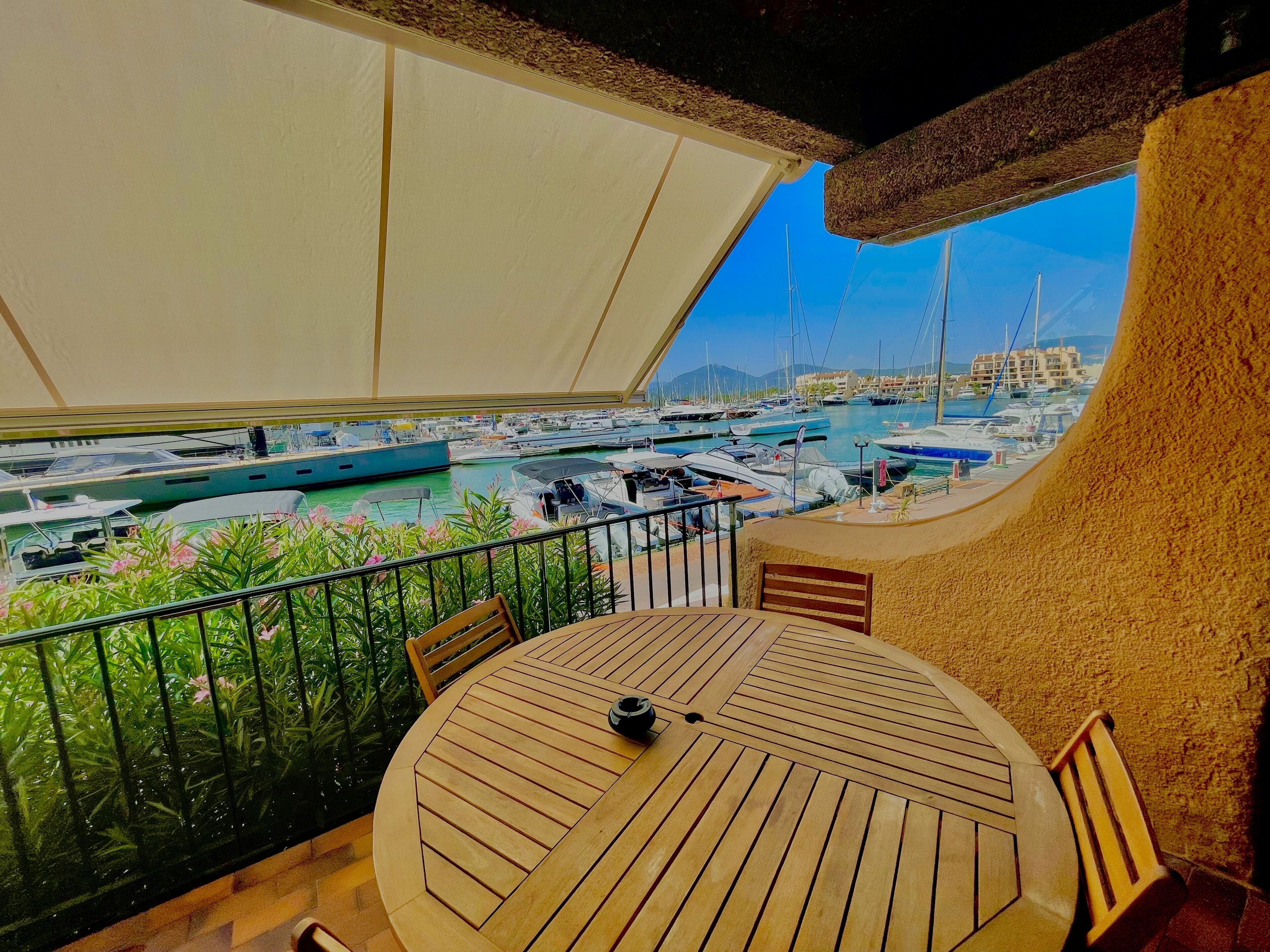 Una mesa redonda de madera con sillas, un dosel blanco y vista a un puerto deportivo con barcos.