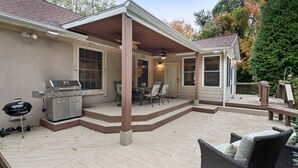 En overdækket terrasse med grill, udendørs møbler og udsigt til huset.