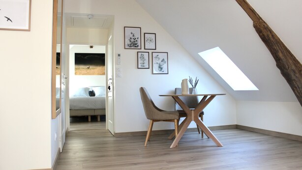 Ein Dachzimmer mit schrägen Wänden, Holzbalken, einem Esstisch mit Stühlen und gerahmten Pflanzenabbildungen an der Wand.