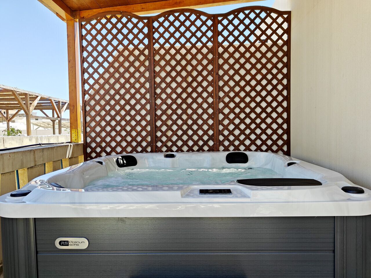 A hot tub with a wooden lattice backdrop, a wooden deck, and a clear sky.