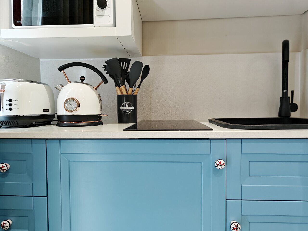 Una cocina con un microondas blanco, una tetera y un tostador sobre la encimera. Hay un recipiente negro que contiene utensilios de cocina. Los armarios son azules con pomos plateados.