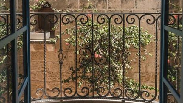 A view through a metal-framed window of a stone building with a balcony. - Zebbuga guest house Malta 5 (Ħaż-Żebbuġ)