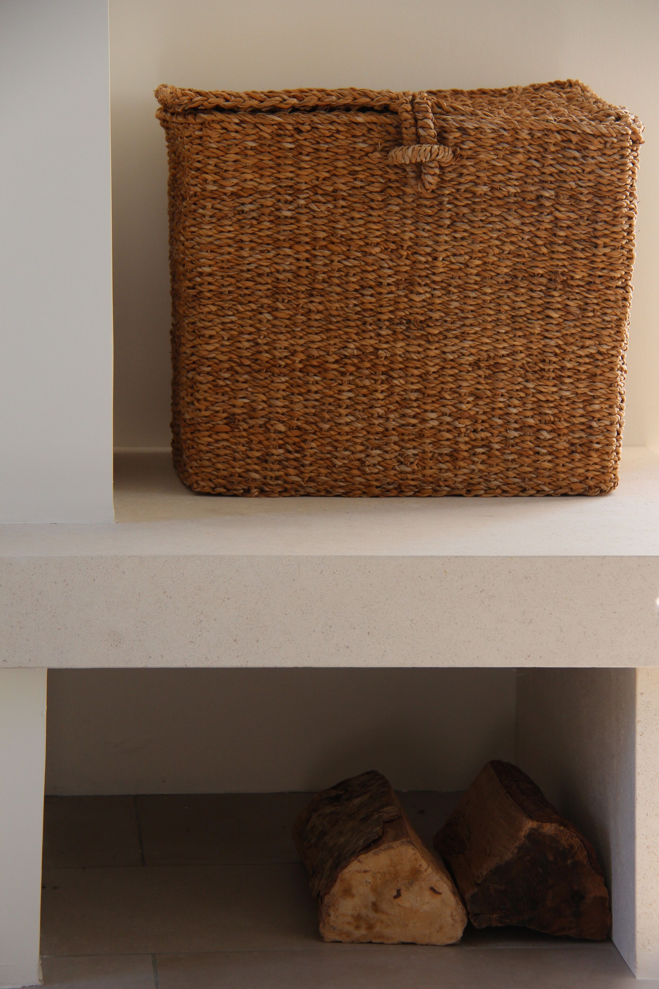 Un panier tressé est posé sur une étagère au-dessus d’une cheminée, avec deux bûches en dessous.