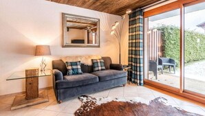 A cozy living room with a dark sofa, a glass coffee table, a wooden side table, a mirror, and a large window with curtains. - 250m from ski lifts, Ground floor, terrace, parking, tv, ski locker, 58m², Morzine (Morzine)