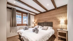 Een slaapkamer met een houten plafond, een groot bed met witte lakens, een nachtkastje met een lamp en een raam met gordijnen.