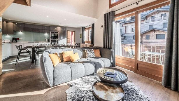 Een moderne woonkamer met een grijze bank, een bijzettafeltje en uitzicht op een balkon met houten gebouwen.