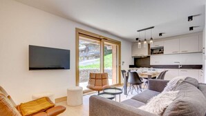 A modern living room with a grey sofa, a brown leather chair, and a flat-screen TV mounted on the wall. There is a dining area with a table and chairs, and a kitchen with white cabinets and pendant lights. - 200m from ski slopes, Ground floor, terrace, tv, 42m², Morzine (Morzine)