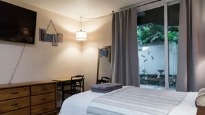Ein Schlafzimmer mit einem Bett, einem Schreibtisch und einem Stuhl. Ein Schiebeglastür führt zu einem Außenbereich mit Zaun und Bäumen.