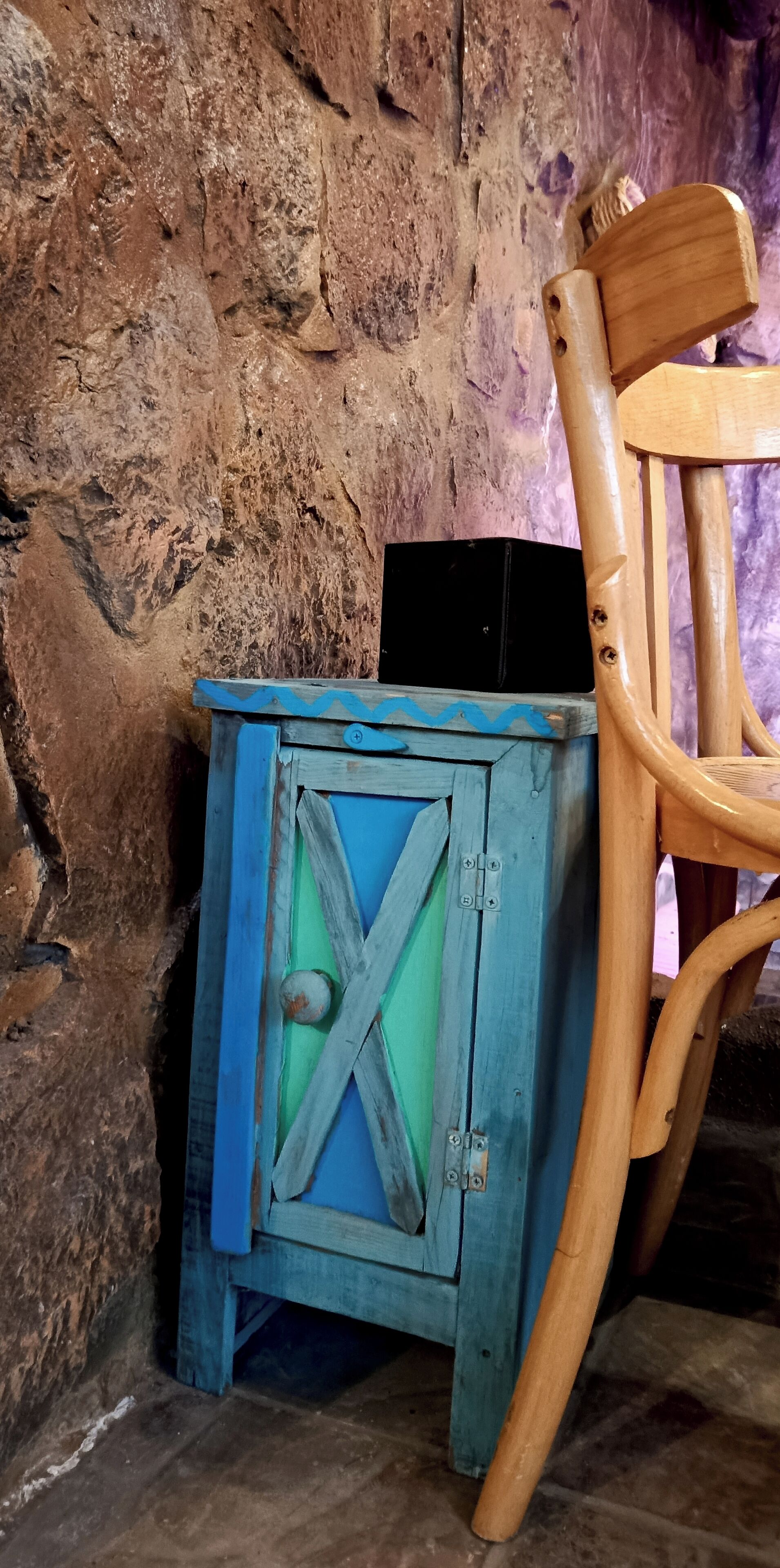 Uma cadeira de madeira rústica e um pequeno armário azul com fecho de metal em frente a uma parede de pedra.