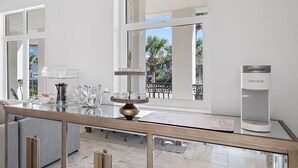 A modern, well-lit room with a glass coffee table, a sleek KEURIG coffee machine, and a view of a balcony with palm trees. - Private Heated Pool - Golf Cart Included (Destin)