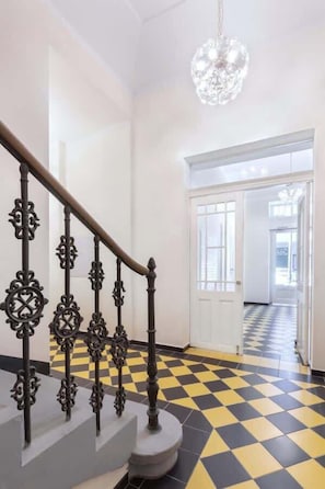 Un escalier orné de rampes en métal ouvragé et surmonté d’un lustre, menant à une pièce dont le sol est à carreaux et où l’on aperçoit une porte au fond.