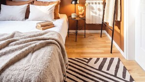 A bedroom with a sloped ceiling, a bed with white and beige bedding, a side table with a lamp, a radiator, and a door with a towel rack.