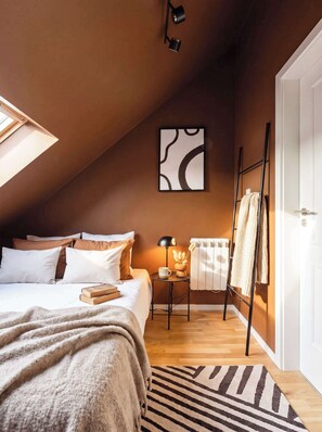 Ein Schlafzimmer mit schrägverdachter Decke, einem Bett mit weißer und beiger Bettwäsche, einem Nachttisch mit Lampe, einem Heizkörper und einer Tür mit Handtuchhalter.