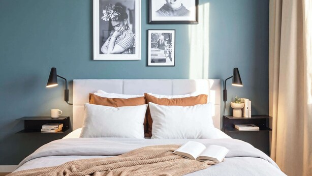 Un lit bien fait, avec des oreillers blancs et bruns, une couverture tricotée et un livre ouvert sur la table de chevet. Trois photographies encadrées sont accrochées au mur, au-dessus du lit.