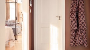 Un couloir avec une porte blanche, un porte-manteau sur lequel est accroché un manteau à motifs, et un aperçu d’une chambre avec un lit et des rideaux.
