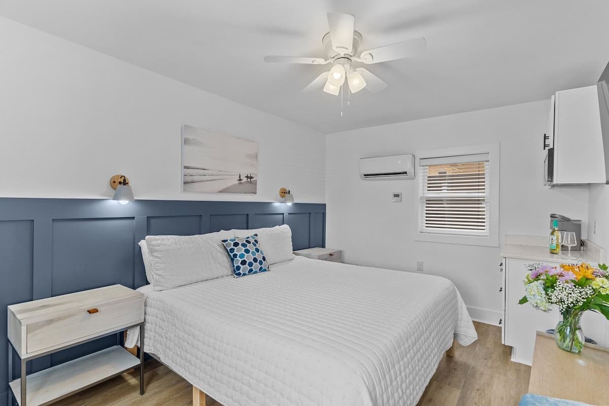 A bedroom with a bed, bedside tables, a ceiling fan, an air conditioning unit, a window with blinds, and a small desk with a vase of flowers.