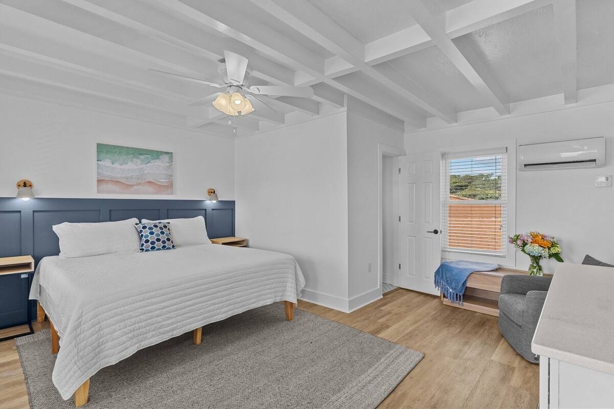 A bedroom with a bed, bedside tables, a nightstand, a ceiling fan, a window with blinds, a door, and a small table with a vase of flowers.