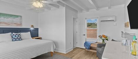 A bedroom with a bed, a nightstand, a desk, and a chair. There is a door leading to a balcony.