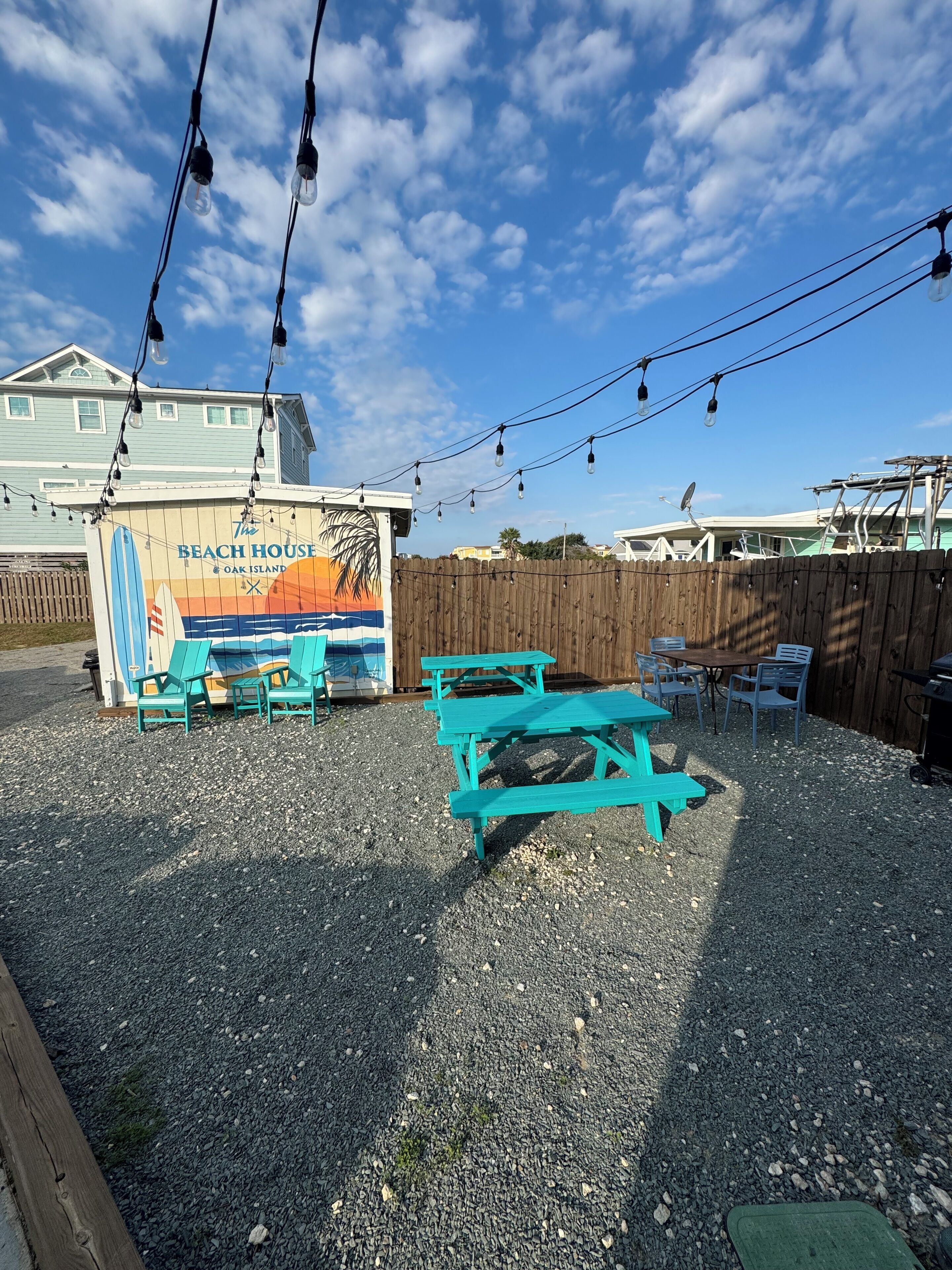 A beach-themed outdoor seating area with turquoise benches and tables, a wooden fence, and a sign for 'The Beach House Oak Island'.
