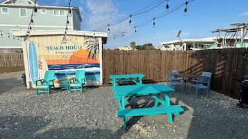 A beach-themed outdoor seating area with turquoise benches and tables, a wooden fence, and a sign for 'The Beach House Oak Island'.
