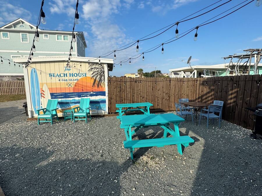 A beach-themed outdoor seating area with turquoise benches and tables, a wooden fence, and a sign for 'The Beach House Oak Island'.
