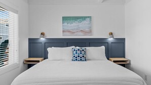 A neatly made bed with a white quilt, two pillows, and a blue patterned pillow. There is a painting of a beach scene above the bed.