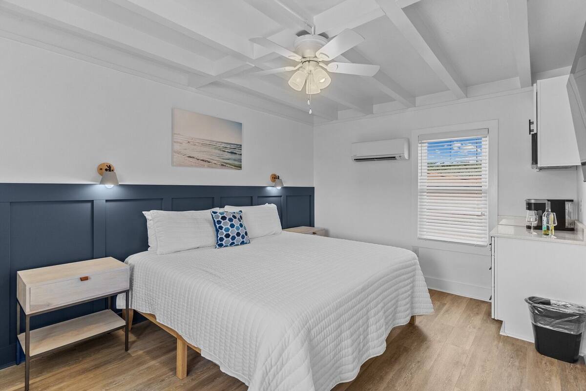 A bedroom with a bed, bedside tables, a ceiling fan, a window with blinds, and a wall-mounted air conditioning unit.