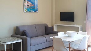 A living room with a grey sofa, a small white table, a white dining table with chairs, a painting of a coastal scene, a flat-screen TV, and an air conditioning unit. - La Finestra sul mare Sorrento (Piano di Sorrento)