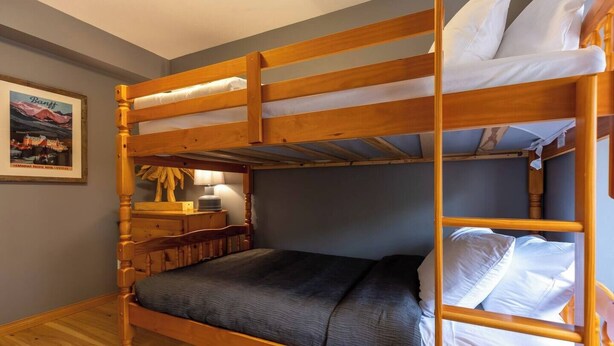 Una cama litera con estructura de madera, colchón gris y almohadas blancas. Hay una mesita de noche de madera con una lámpara y un cuadro en la pared.