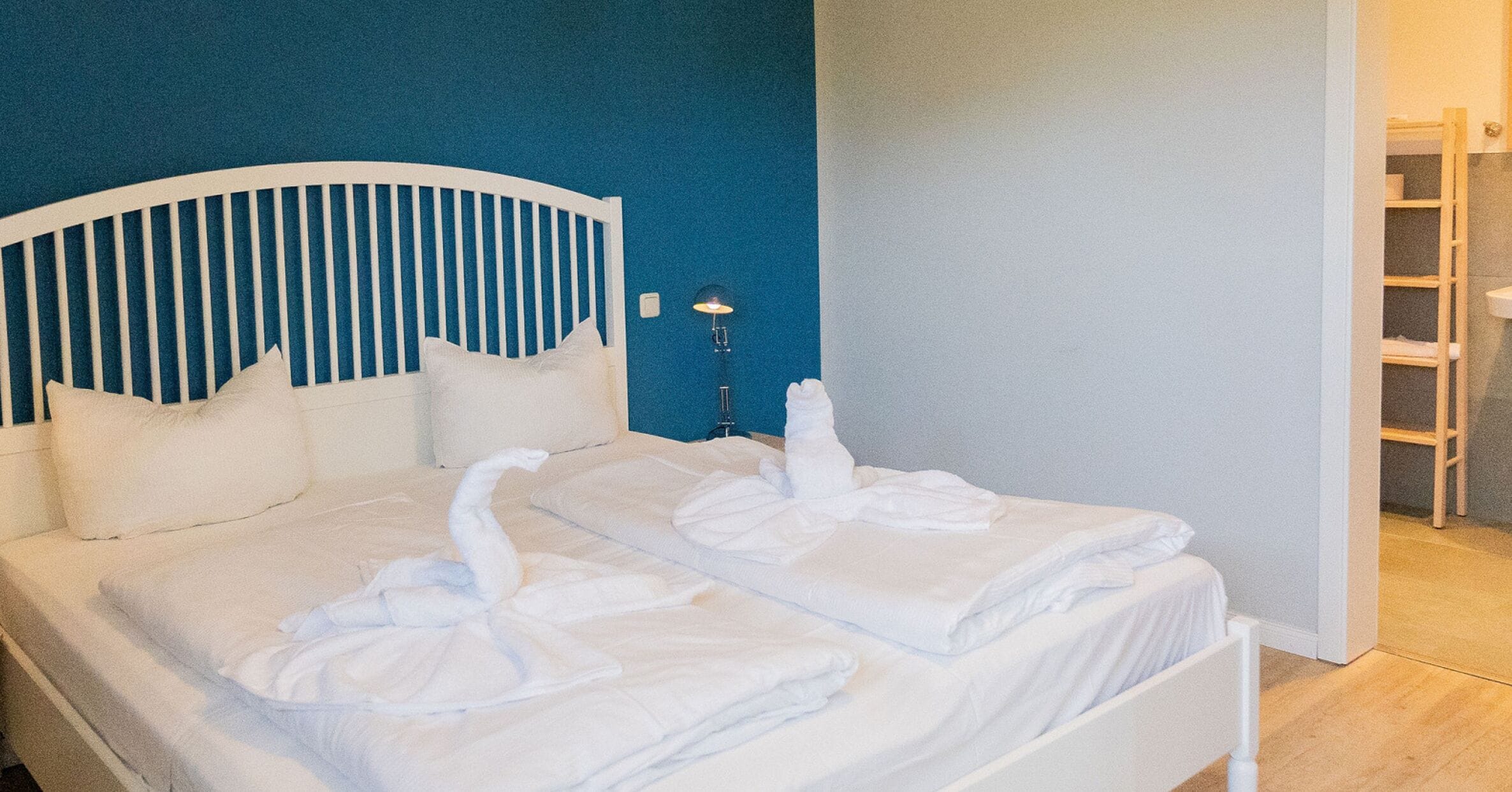 A neatly made bed with white linens and towels arranged in a swan shape, a white headboard, and a bedside lamp.