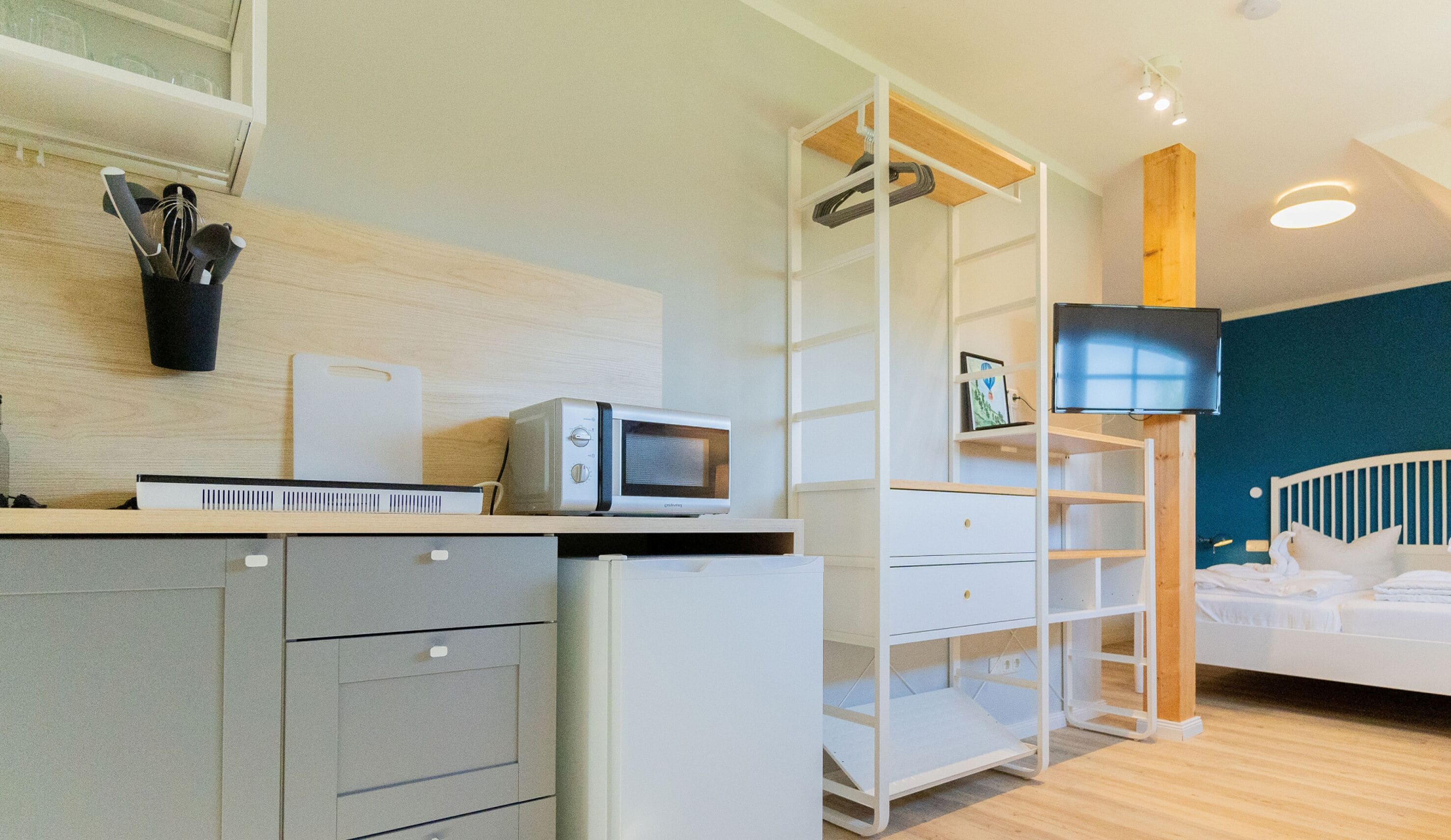 A modern hotel room with a kitchenette, a flat-screen TV, a bed, and a wooden ceiling.