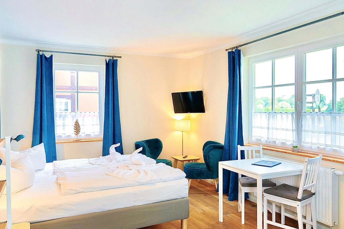 A hotel room with a bed, a desk, two chairs, a TV, and a window with curtains.