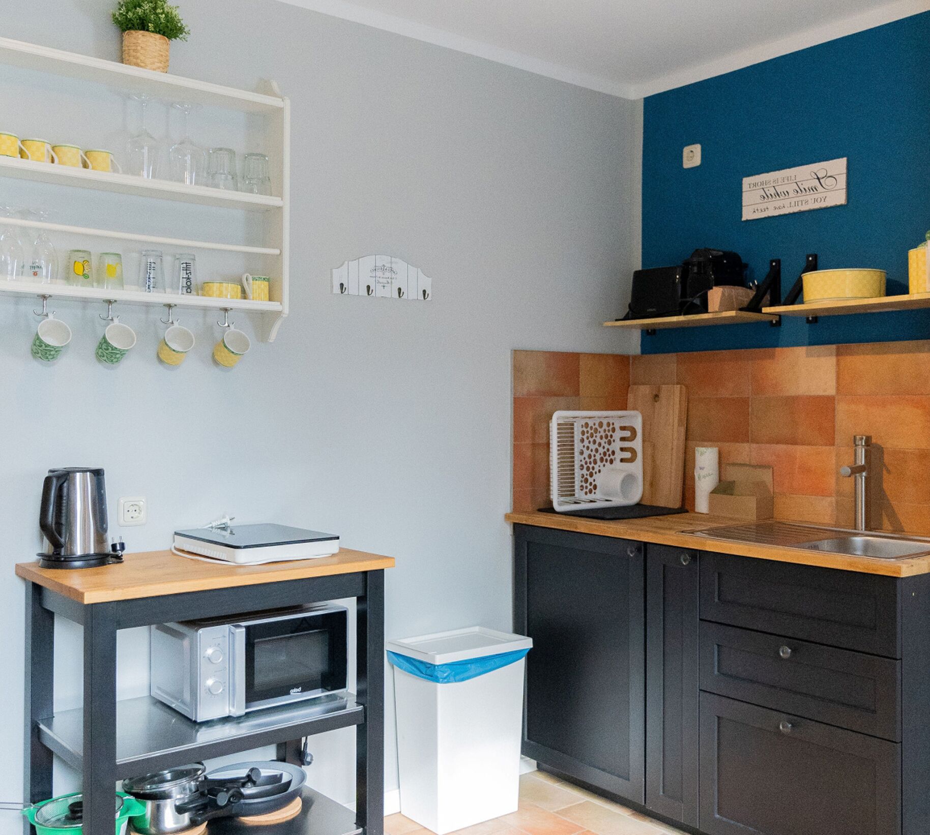 Cocina compacta con microondas, hervidor de agua, un lavamanos pequeño y estantes con cristalería y una cafetera.