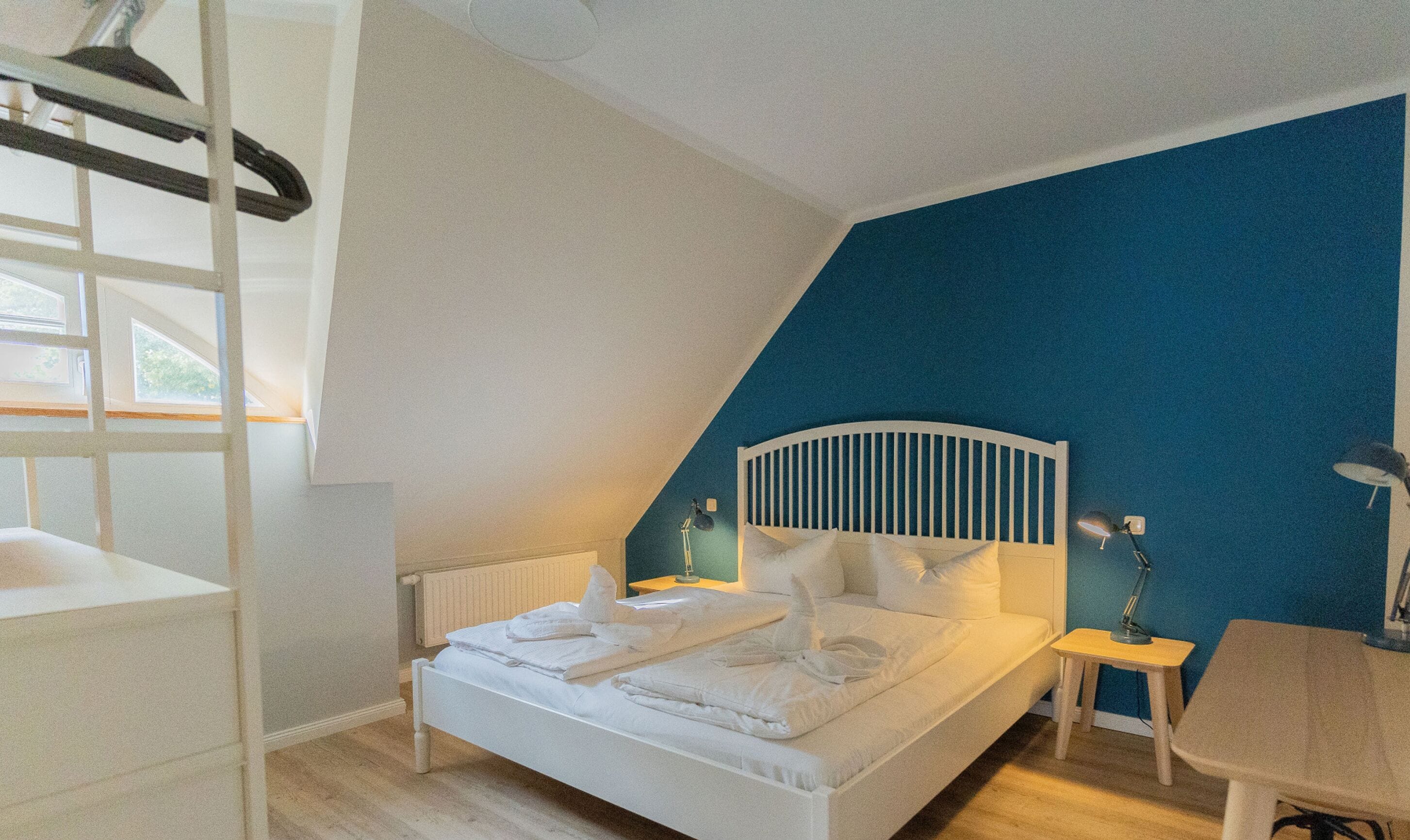 Ein Schlafzimmer mit einem weißen Bett, einer blauen Akzentwand, Holzboden und einem Fenster mit Blick.